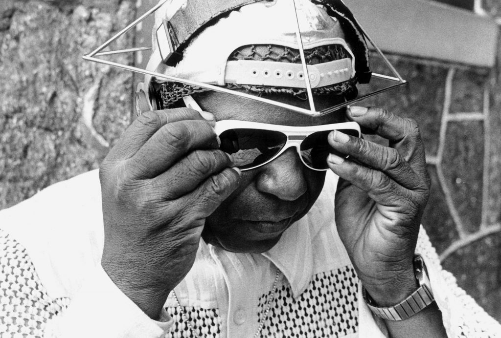 Black-and-white photo of Sun-ra adjusting sunglasses while wearing a geometric wire headpiece over a cap.