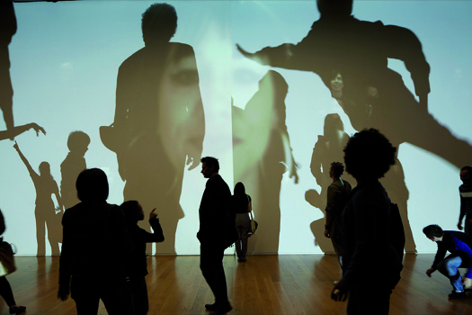 People interact with People on People (2010) by Rafael Lozano-Hemmer, an interactive installation projecting enlarged, shadow-like silhouettes of human figures onto a wall as visitors move through the space.