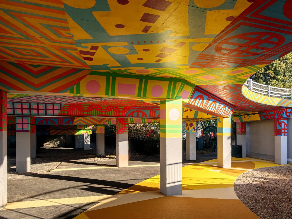 A vibrant public space underneath a concrete overpass covered in colourful geometric murals.